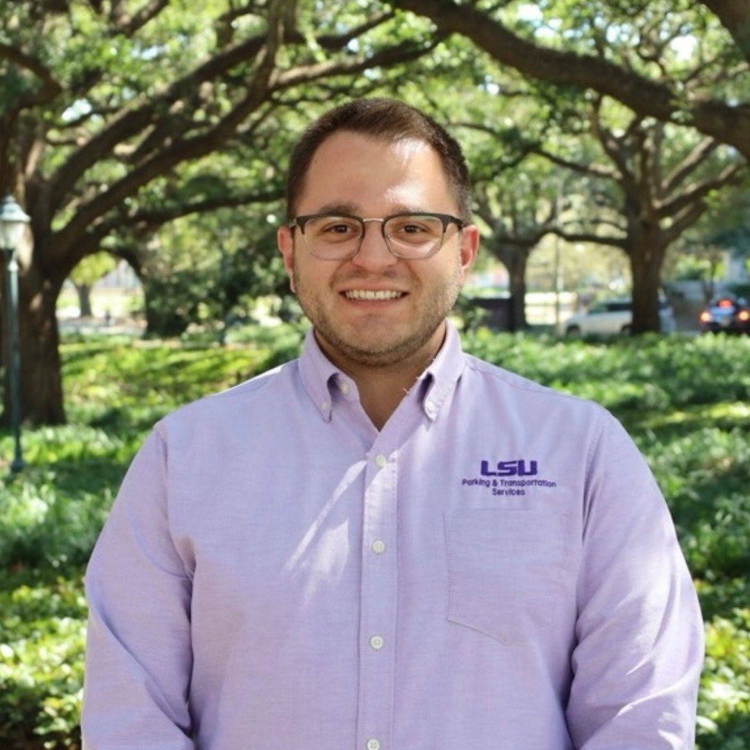 Man in purple shirt smiling and standing in front of a tree and bushes